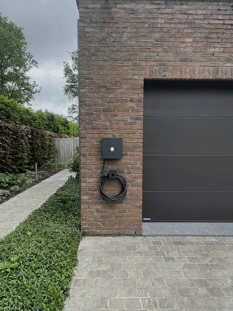 Zwarte laadpaal op bakstenen gevel naast garagepoort, mooie oprit, geplaatst door Wattra