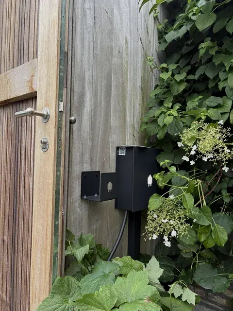 Zwarte Smappee laadpaal in groene tuin naast houten deur, geplaatst door Wattra