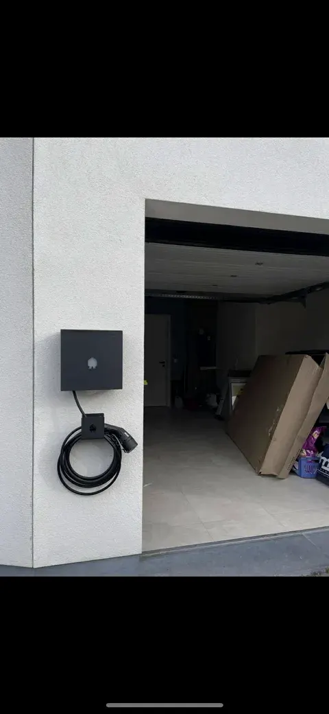 Zwarte laadpaal aan witte gevel naast open garagepoort, geplaatst door Wattra